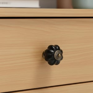 May include: Close-up of a light wood drawer with a black, flower-shaped knob. The knob has a bronze-colored base and a detailed, sunburst-like center. The drawer's wood grain is visible, and the overall aesthetic is simple and elegant.