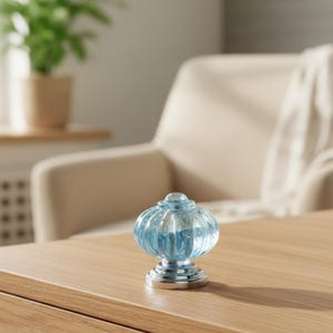 May include: A decorative blue glass knob with a ribbed, pumpkin-shaped design. The knob sits on a silver-colored base. The knob is on a wooden surface.