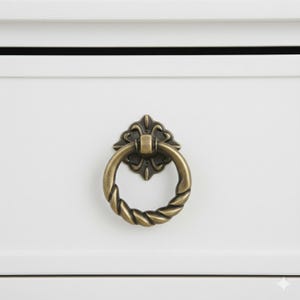 May include: Close-up of a bronze-colored drawer pull with a twisted ring handle and ornate backplate. The hardware is mounted on a white drawer front, showcasing a classic design element for furniture.