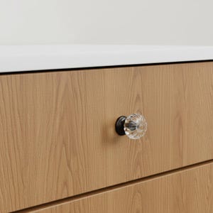 May include: A clear, ribbed glass cabinet knob with a black metal base is attached to a light brown wooden drawer. The drawer is part of a larger cabinet or piece of furniture. The background is white.