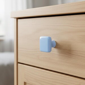 May include: Close-up of a light wood dresser drawer with a square, light blue knob. The knob is centered on the drawer, providing a simple, modern aesthetic. The wood grain is visible, adding texture to the piece.