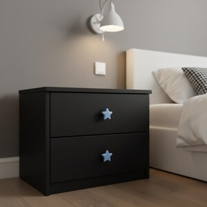 Puede incluir: Una mesita de noche negra de dos cajones con tiradores en forma de estrella azul claro. La mesita de noche est&aacute; colocada junto a una cama con ropa de cama blanca y una almohada con estampado en blanco y negro. Una l&aacute;mpara de pared blanca est&aacute; encima.