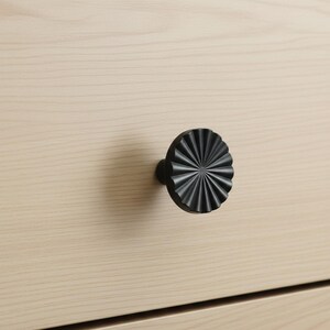 May include: Close-up of a black, decorative cabinet knob with a radial design. The knob is mounted on a light-colored wooden drawer front. The knob's design features a series of radiating lines, creating a textured, starburst appearance. The hardware adds a modern touch.