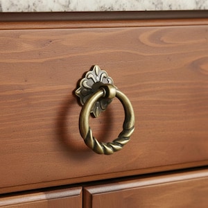 May include: Antique brass drawer pull with a decorative floral backplate and a twisted ring handle. The hardware is mounted on a stained wooden cabinet. The drawer pull is a classic design element for furniture.