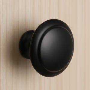 May include: A close-up of a black cabinet knob with a rounded design. The knob is mounted on a light-colored wooden surface. The knob has a matte finish and a simple, classic style.