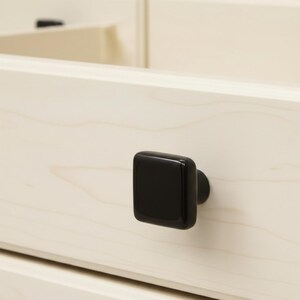 May include: A close-up of a light-colored wooden drawer with a square, black cabinet knob. The knob is centered on the drawer, and the wood grain is visible. The knob has a glossy finish.