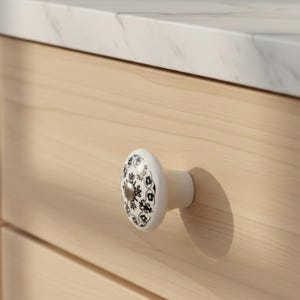 May include: A white ceramic cabinet knob with a black floral pattern and a silver center. The knob is attached to a light-colored wooden drawer. The countertop above is white with gray veining.