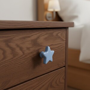 Puede incluir: Una mesita de noche de madera con un tirador en forma de estrella azul claro. La mesita tiene un acabado marr&oacute;n c&aacute;lido y est&aacute; junto a una cama con una almohada blanca. El tirador en forma de estrella a&ntilde;ade un toque caprichoso.