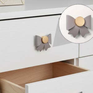 May include: Gray bow-shaped drawer pulls with gold-colored circular centers. The drawer pulls are attached to a white dresser. The drawer is open, revealing the wooden interior.