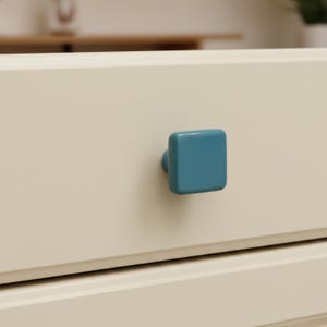 May include: Close-up of a cream-colored drawer with a square, teal-colored knob. The knob is centered on the drawer front, providing a simple, modern aesthetic. The drawer appears to be part of a larger piece of furniture.