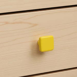 May include: Close-up of a light-colored wooden drawer with a bright yellow square knob. The knob is centered on the drawer front, providing a pop of color against the wood grain.