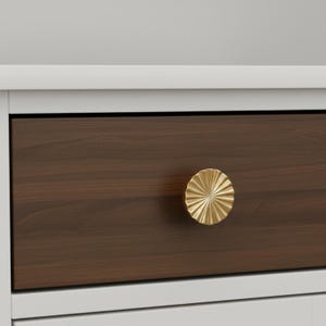 May include: Close-up of a drawer with a dark brown wood front and a gold-colored, sunburst-style knob. The drawer is set within a white frame, suggesting a piece of furniture like a dresser or cabinet.