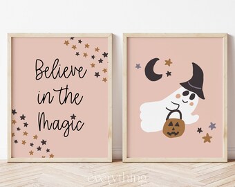 May include: Two pink framed prints with a Halloween theme. The first print has the text "Believe in the Magic" with gold and black stars. The second print has a white ghost wearing a witch hat and holding a pumpkin bucket. The ghost is flying with a crescent moon in the background.