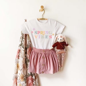 Pu&ograve; includere: Maglietta bianca con una grafica colorata "Spring Vibes" e un design floreale. La maglietta &egrave; appesa a un appendiabiti di legno. Sono anche raffigurati una gonna di velluto a coste rosa e un coniglio di peluche.