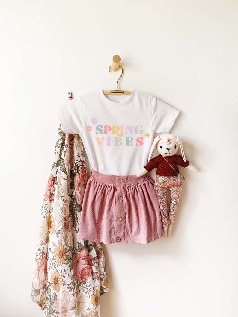 Pu&ograve; includere: Maglietta bianca con una grafica colorata "Spring Vibes" e un design floreale. La maglietta &egrave; appesa a un appendiabiti di legno. Sono anche raffigurati una gonna di velluto a coste rosa e un coniglio di peluche.