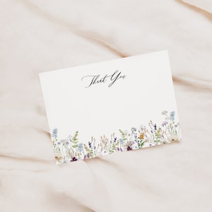 Op de afbeelding: Een witte bedankkaart met een bloemige rand. De bloemen zijn in tinten paars, blauw, geel en groen. De tekst "Bedankt" staat in zwarte cursieve letters.