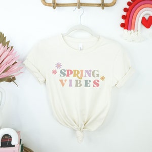 Pu&ograve; includere: Una maglietta bianca con il testo "SPRING VIBES" in lettere colorate e fiori su entrambi i lati del testo.