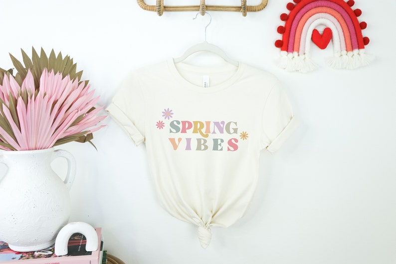 Pu&ograve; includere: Una maglietta bianca con il testo "SPRING VIBES" in lettere colorate e fiori su entrambi i lati del testo.