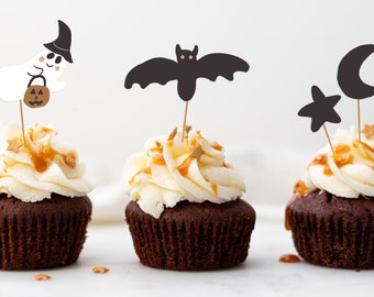 Peut inclure: Trois cupcakes au chocolat surmontés d'un glaçage blanc et d'un filet de caramel. Chaque cupcake est décoré d'un topper sur le thème d'Halloween : un fantôme, une chauve-souris et un croissant de lune avec une étoile.