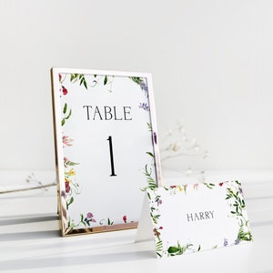 May include: A white table number card with a floral border in shades of pink, purple, and green. The card is framed in gold and reads "TABLE 1". A white place card with a similar floral border reads "HARRY".