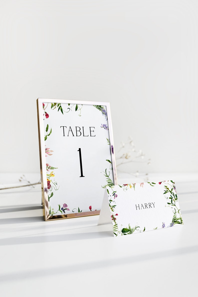 May include: A white table number card with a floral border in shades of pink, purple, and green. The card is framed in gold and reads "TABLE 1". A white place card with a similar floral border reads "HARRY".