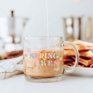 Pu&ograve; includere: Una tazza di vetro con manico piena di caff&egrave; e panna. La tazza ha il testo "SPRING VIBES" in lettere colorate con fiori su entrambi i lati.