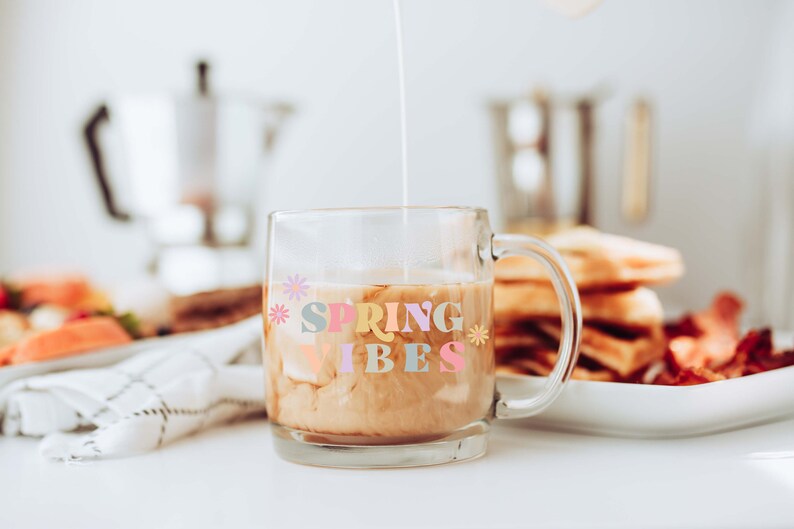 Pu&ograve; includere: Una tazza di vetro con manico piena di caff&egrave; e panna. La tazza ha il testo "SPRING VIBES" in lettere colorate con fiori su entrambi i lati.