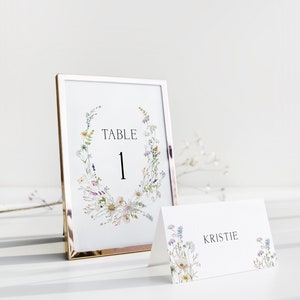 Op de afbeelding: Een witte tafelnummerkaart met een gouden frame en een bloemenontwerp. De kaart zegt "Tafel 1". Een witte plaatskaart met een bloemenontwerp zegt "Kristie".