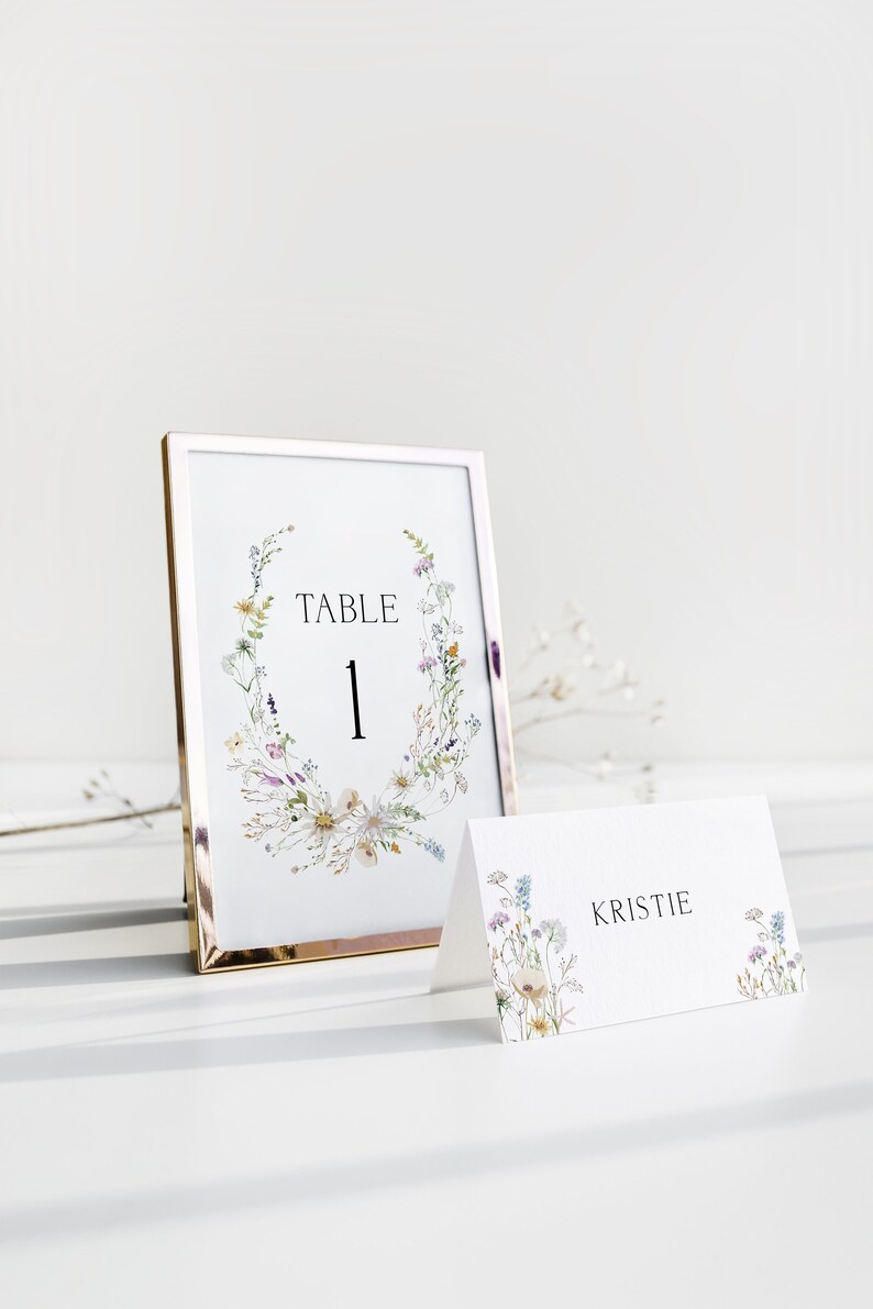 Op de afbeelding: Een witte tafelnummerkaart met een gouden frame en een bloemenontwerp. De kaart zegt "Tafel 1". Een witte plaatskaart met een bloemenontwerp zegt "Kristie".