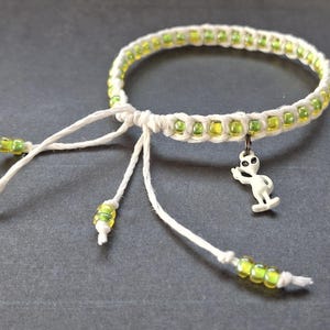 May include: A handmade macrame bracelet with a white cord, accented with yellow and green beads. The bracelet features a small white alien charm. The adjustable ties also have matching beads.