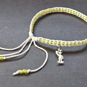 May include: A handmade bracelet featuring a white woven band with yellow-green beads. The bracelet has a small alien charm and white string tassels with matching beads. The bracelet is on a dark gray surface.