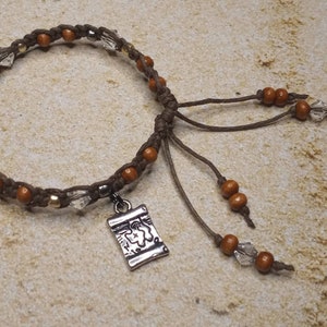 May include: A brown and gold beaded bracelet with a silver charm featuring a map design. The bracelet has a brown cord with wooden beads and crystal beads.