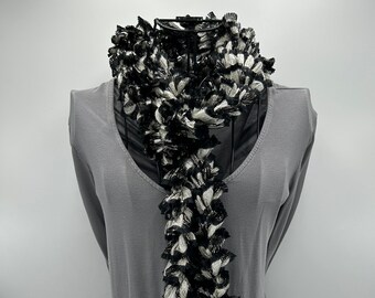 May include: A black and white frilled scarf on a grey mannequin. The scarf features a textured, layered design with alternating black and white stripes. The scarf is draped over a grey top.