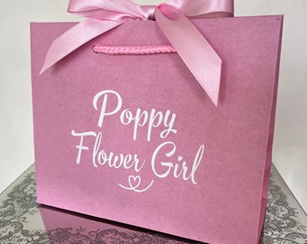May include: A pink gift bag with the words "Poppy Flower Girl" in white script. The bag has a pink ribbon tied in a bow at the top and pink rope handles. The bag is sitting on a mirrored surface.
