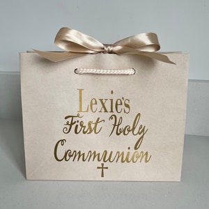Pode incluir: Uma sacola de presente bege com um la&ccedil;o de cetim dourado e al&ccedil;as de corda. A sacola apresenta o texto dourado "Lexie's First Holy Communion" com um s&iacute;mbolo de cruz abaixo. A sacola &eacute; projetada para uma celebra&ccedil;&atilde;o religiosa.
