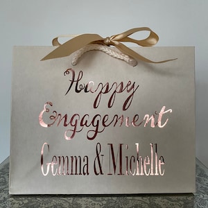 May include: A cream-colored gift bag with the words "Happy Engagement Gemma & Michelle" in rose gold script. The bag has a gold ribbon and rope handles, perfect for a special occasion.