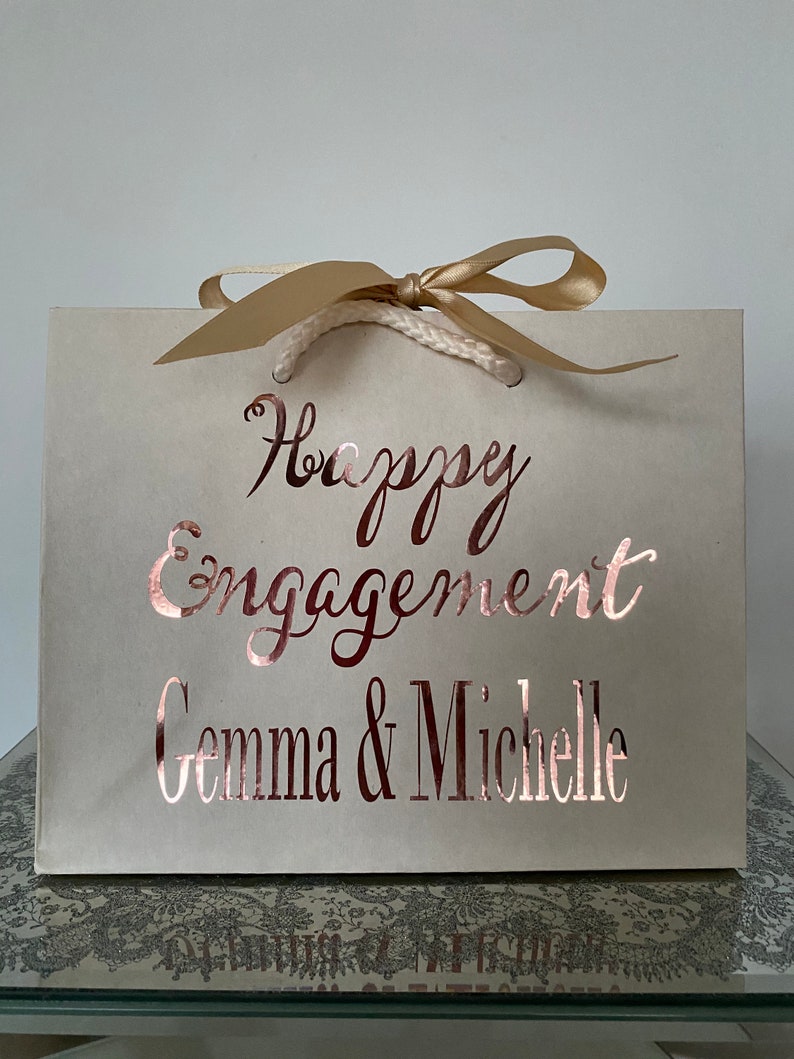 May include: A cream-colored gift bag with the words "Happy Engagement Gemma & Michelle" in rose gold script. The bag has a gold ribbon and rope handles, perfect for a special occasion.