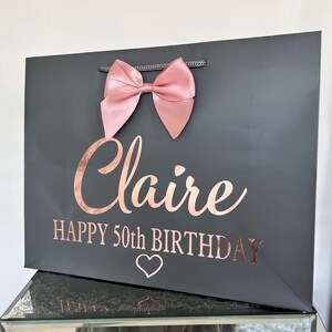 May include: A dark gray gift bag with a large pink bow at the top. The bag has the name "Claire" and "HAPPY 50th BIRTHDAY" in rose gold lettering, along with a heart symbol.
