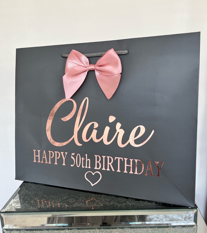 May include: A dark gray gift bag with a large pink bow at the top. The bag has the name "Claire" and "HAPPY 50th BIRTHDAY" in rose gold lettering, along with a heart symbol.