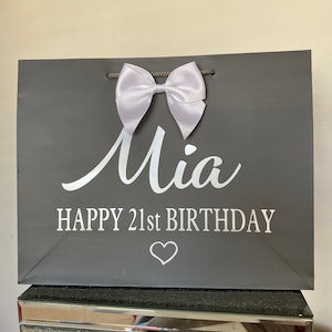 May include: A gray gift bag with a white satin bow and the name "Mia" in cursive. The text "HAPPY 21st BIRTHDAY" and a heart are printed below. The bag has gray rope handles.