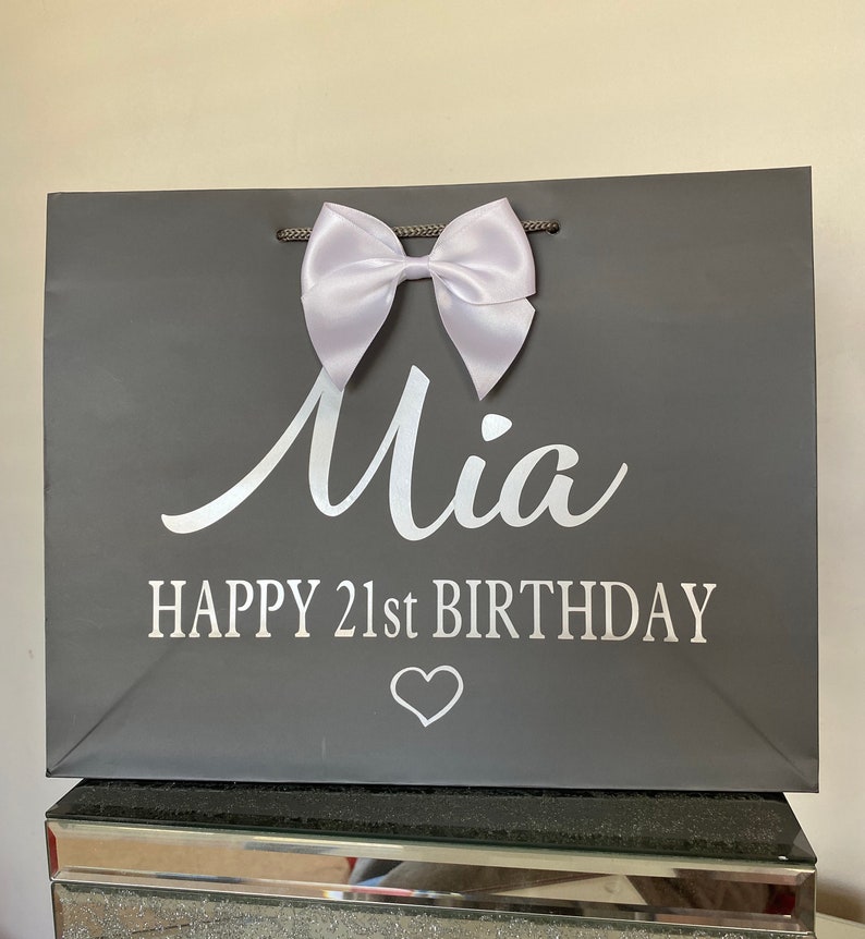 May include: A gray gift bag with a white satin bow and the name "Mia" in cursive. The text "HAPPY 21st BIRTHDAY" and a heart are printed below. The bag has gray rope handles.