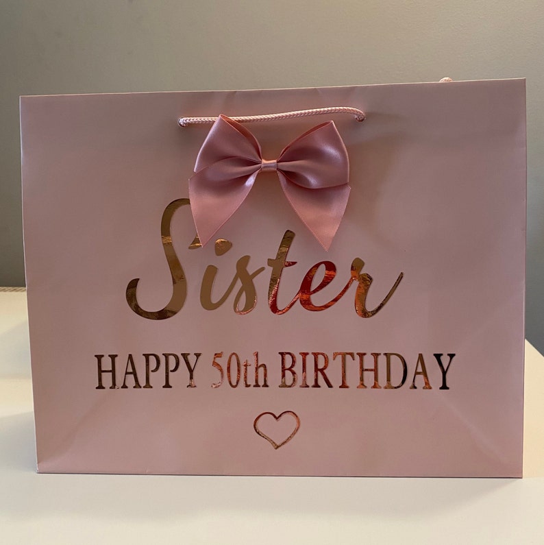 May include: A pink gift bag with a satin bow and rope handles. The words "Sister" and "HAPPY 50th BIRTHDAY" are printed in gold. A small heart is printed below the text.