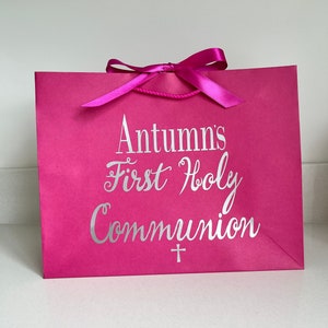 Pode incluir: Uma sacola de presente rosa brilhante com uma fita e al&ccedil;as de corda combinando. A sacola apresenta o texto "Autumn's First Holy Communion" em prata, junto com um pequeno s&iacute;mbolo de cruz. Um item festivo e comemorativo.