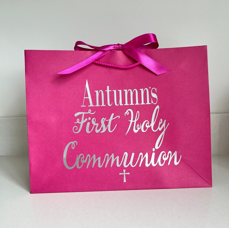 Pode incluir: Uma sacola de presente rosa brilhante com uma fita e al&ccedil;as de corda combinando. A sacola apresenta o texto "Autumn's First Holy Communion" em prata, junto com um pequeno s&iacute;mbolo de cruz. Um item festivo e comemorativo.