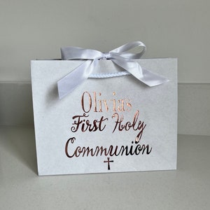 Pode incluir: Uma sacola de presente branca com um la&ccedil;o de fita branco e uma al&ccedil;a de cord&atilde;o branca. A sacola tem o texto "Olivia's First Holy Communion" em escrita cor de cobre, com uma pequena cruz abaixo.