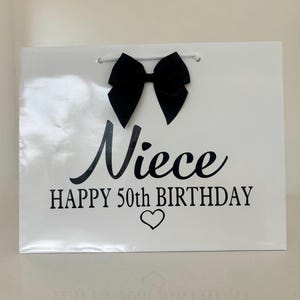 May include: A white gift bag with a black bow and the words "Niece HAPPY 50th BIRTHDAY" in black script. A small heart is below the text. The bag has a white cord handle.