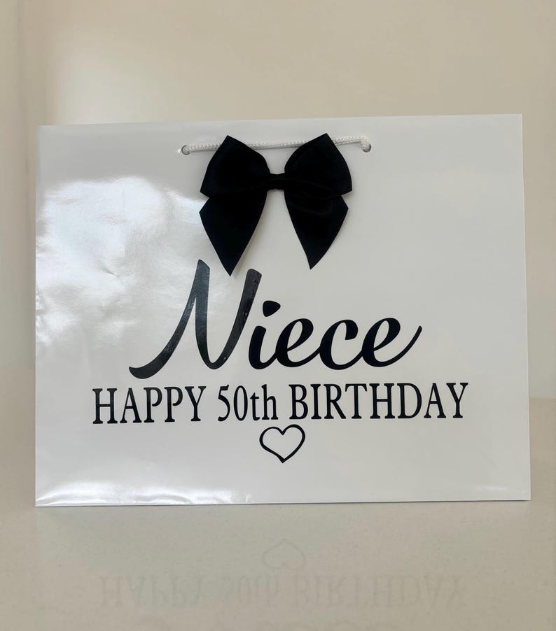 May include: A white gift bag with a black bow and the words "Niece HAPPY 50th BIRTHDAY" in black script. A small heart is below the text. The bag has a white cord handle.