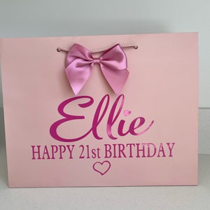 May include: A pink gift bag with a large pink bow at the top. The bag has the name "Ellie" and the text "HAPPY 21st BIRTHDAY" in a darker pink font, along with a heart symbol.
