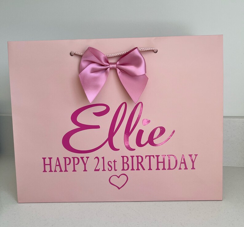May include: A pink gift bag with a large pink bow at the top. The bag has the name "Ellie" and the text "HAPPY 21st BIRTHDAY" in a darker pink font, along with a heart symbol.
