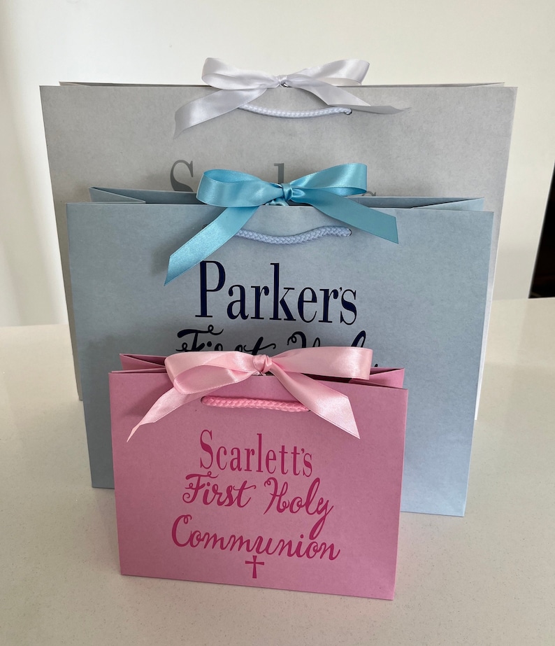 Pode incluir: Tr&ecirc;s sacolas de presente em tamanhos variados. A maior &eacute; cinza, a do meio azul claro e a menor rosa. Cada sacola tem um la&ccedil;o de fita combinando. A sacola azul clara tem o nome "Parkers" impresso. A sacola rosa diz "Scarlett's First Holy Communion".